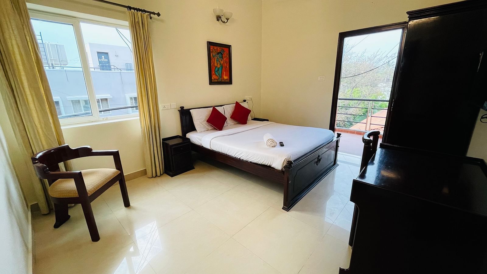 A well-lit room showcasing a double bed with white linens and red cushions, an armchair, a window with natural light, and a doorway leading to the balcony at Sanctum Suites Indiranagar, Bangalore.