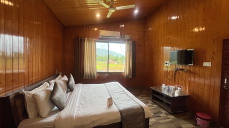Side view of Daksh Elite Cottages showcasing it's double bed facing a wall-mounted TV, and a window beside offering views of the surroundings.
