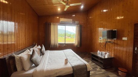 Side view of Daksh Elite Cottages showcasing it's double bed facing a wall-mounted TV, and a window beside offering views of the surroundings.