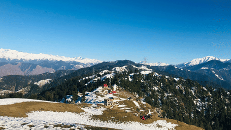 A snow-covered mountain slope with patches of green grass under a clear blue sky.