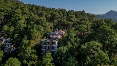 A drone shot taken during the day of DLS Whispering Winds, Kasauli, surrounded by lush greenery and the backdrop of a clear sky.