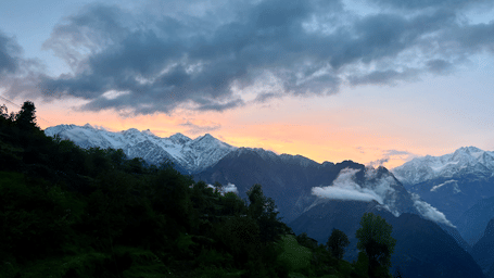 Mountain landscape at Faraway Cottages, Auli, showing forested hills and snow-capped peaks under a dramatic sunset sky. A trip to Auli from Mumbai is 1,800 km by road.