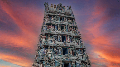 An intricate, multi-tiered Dravidian-style Hindu temple gopuram adorned with colorful sculptures of deities against a dramatic sunset sky.