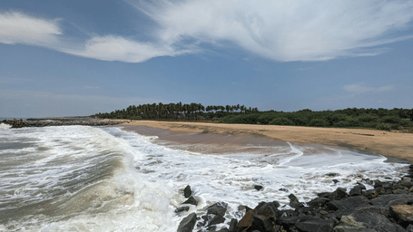 A wide-angle view of a rugged coastline where white ocean waves crash against a dark, rocky shoreline. In the background, a sandy beach transitions into a dense line of green trees under a bright blue sky with wispy white clouds.