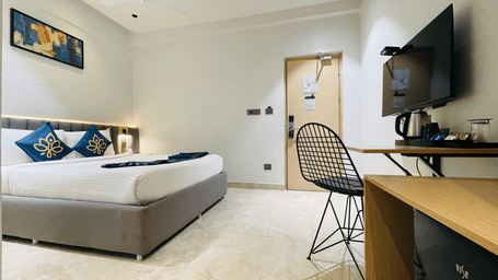 King bed room at Hotel Kyzen HiTech City featuring navy blue accent bedding, wall-mounted TV, framed artwork, wooden work desk, wire chair, and ceiling fan.