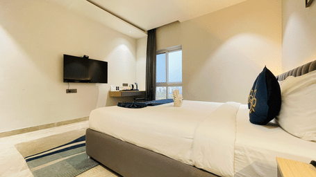 Corner view of a hotel room at Hotel Kyzen HiTech City with a king bed, wall-mounted TV, grey sofa seating, and large window with natural daylight.