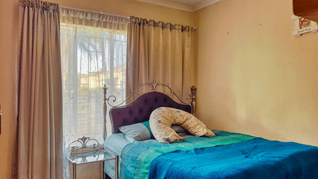 Alt Text: Warm hotel bedroom featuring a bed with bright blue and green bedding, dark purple headboard, plush neck pillow, and sheer window drapes.