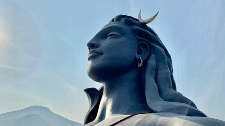 A low-angle profile view of the large black Adiyogi Shiva statue stands against a clear blue sky, surrounded by mist-covered mountains.