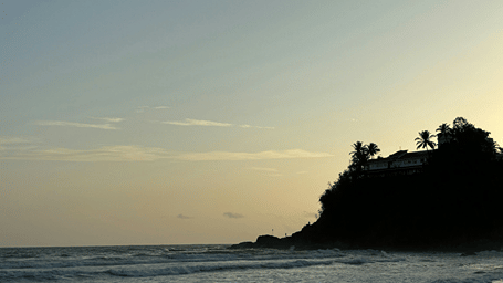 A view of a rocky hill with trees on it and waves crashing on the shore during twilight hour at Baga Beach. It is one of the best places to visit in North Goa.