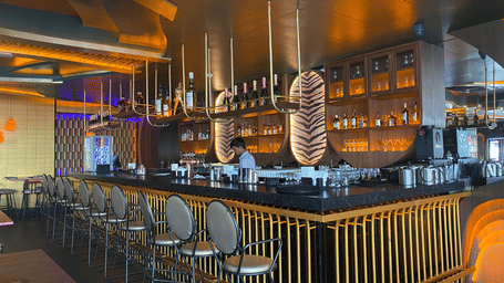 A bar with glasses placed on an elongated slab with illuminated ceiling, and chairs.
