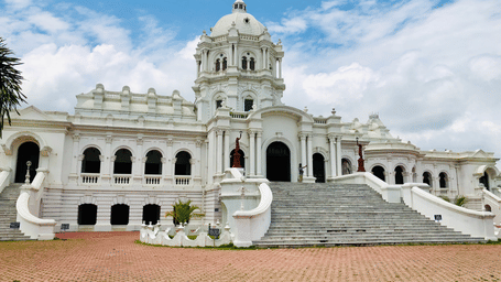 Ujjayanta Palace in Agartala featuring white domed Indo-Saracenic architecture with grand staircase, arched verandas and manicured grounds under blue sky.