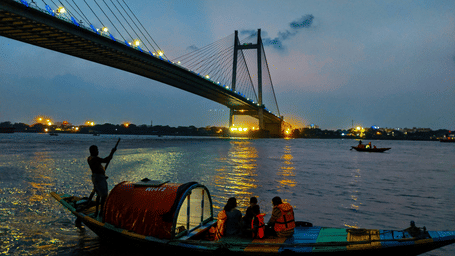 Cable-stayed river bridge lit at dusk with a small wooden ferry boat carrying passengers in life jackets across calm dark water below.