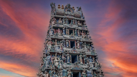 Ornately carved temple gopuram tower with rows of painted deity figures silhouetted against a vivid orange-and-blue sunset sky with drifting clouds.