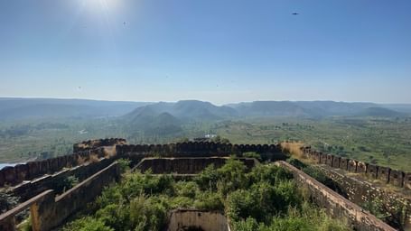 Bright midday shot of the fort’s ruined walls filled with vegetation, overlooking an expansive verdant valley and distant hills under a clear sky, with birds gliding in the sunlight.