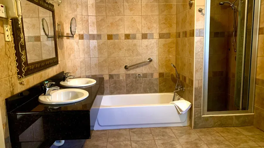 A spacious bathroom with tiled walls and floor includes a bathtub, two sinks with mirrors, and a glass-enclosed shower | Alexander Suite | Altamont Court, Jamaica