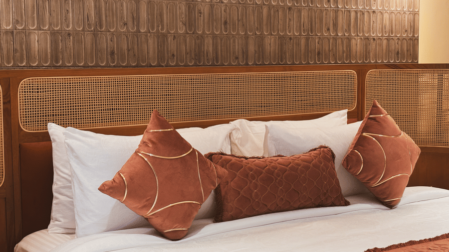 White pillows and 3 brown decorative cushions rest against a light brown woven headboard and a dark textured wall at Avanya Resorts N Retreat.