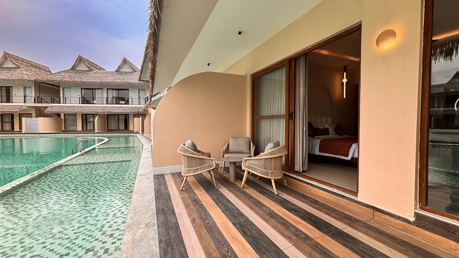 Wicker chairs and a small table sit on a wooden-planked balcony overlooking a swimming pool at Avanya Resorts N Retreat.