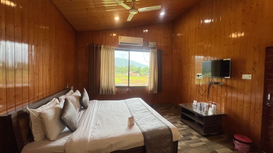 Side view of Daksh Elite Cottages showcasing it's double bed facing a wall-mounted TV, and a window beside offering views of the surroundings.
