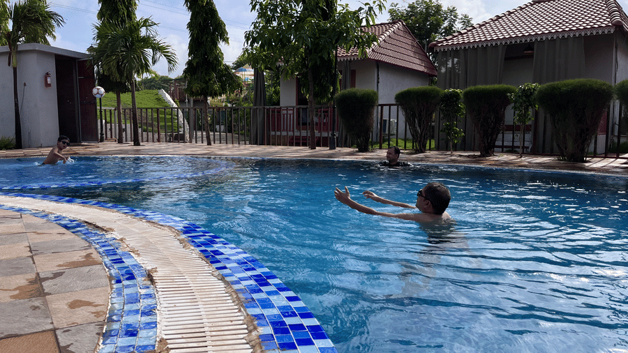 Guests swimming in a large outdoor pool with views of poolside cottages and palm trees - DAN Resorts & Weddings, Palghar