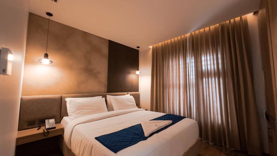 A wide-angle shot of the bedroom at the Apartment Suite featuring a plush double bed with brown curtains covering the windows at F Hotels, Bengaluru.
