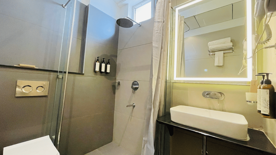 A modern bathroom at Hotel Kyzen HiTech City featuring a glass enclosed shower unit wall-mounted sink with LED mirror dark tiled accents and contemporary fittings in a compact layout.