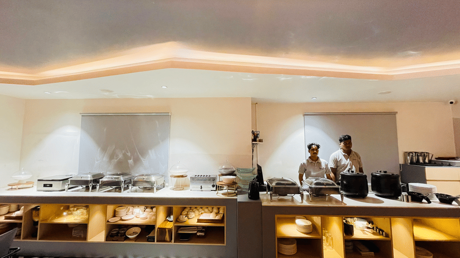 A restaurant buffet area at Hotel Kyzen HiTech City featuring a long counter with food displays staff in white uniform patterned flooring and warm ambient ceiling lighting