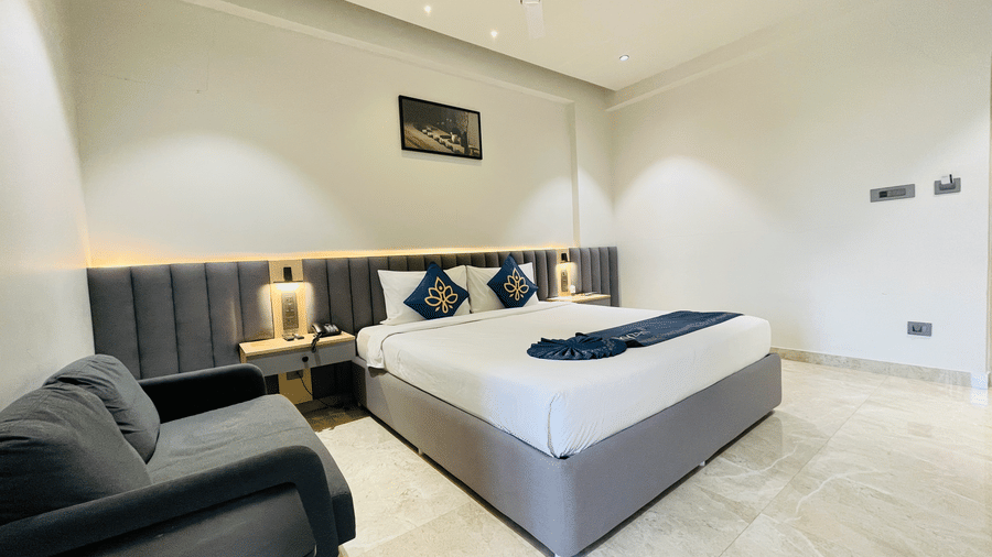A side angle view of a hotel room at Hotel Kyzen HiTech City featuring a king bed with blue accent bedding grey headboard a grey sofa and warm ambient wall lighting on both sides.