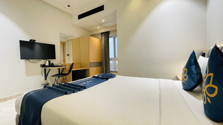 A compact hotel room at Hotel Kyzen HiTech City featuring a king bed with blue accent bedding a wall-mounted TV work desk and modern furnishings in a clean minimal layout.