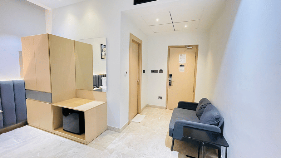 An entrance view of a hotel room at Hotel Kyzen HiTech City featuring a wooden wardrobe blue sofa seating light tiled flooring and a dark entrance door in a modern layout.
