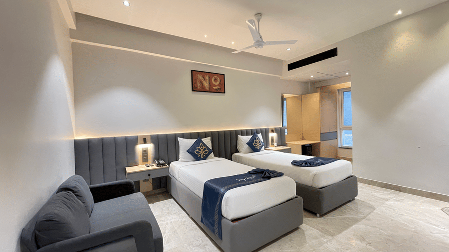 A full interior view of a room at Hotel Kyzen HiTech City featuring two beds with blue accents grey sofa wooden work desk ceiling fan and warm ambient lighting in a modern setting.
