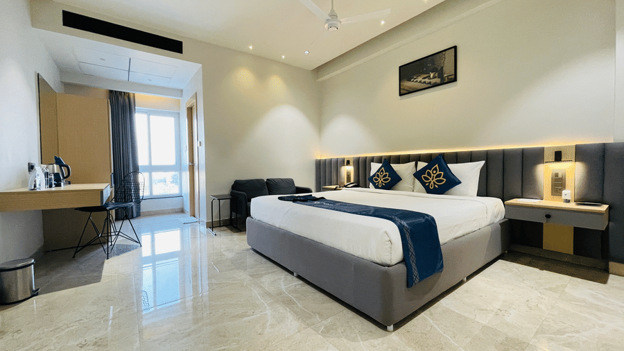 A full layout view of a hotel room at Hotel Kyzen HiTech City featuring a king bed with blue accents grey headboard wooden work desk large windows with blue curtains and light tiled flooring.
