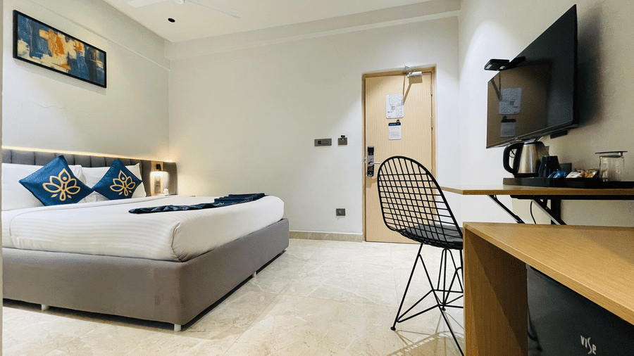 A hotel room at Hotel Kyzen HiTech City featuring a king bed with blue accent pillows wall-mounted TV wooden work desk wire chair and framed artwork on the grey wall.