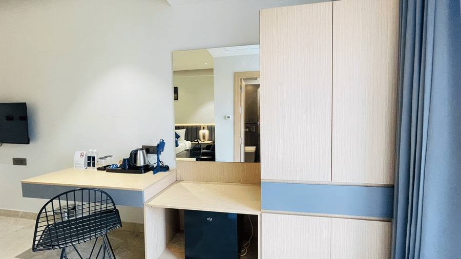 A hotel room corner at Hotel Kyzen HiTech City featuring a wooden wardrobe with mirror a work desk with chair blue curtains and light tiled flooring in a modern layout.