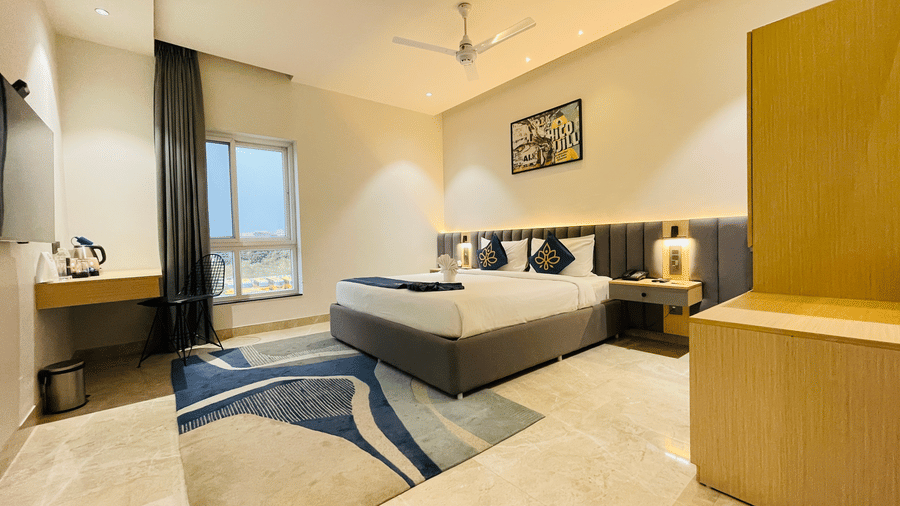 A wide angle view of a room at Hotel Kyzen HiTech City featuring a king bed with blue accent pillows yellow accent wall wooden furniture patterned rug and large windows.