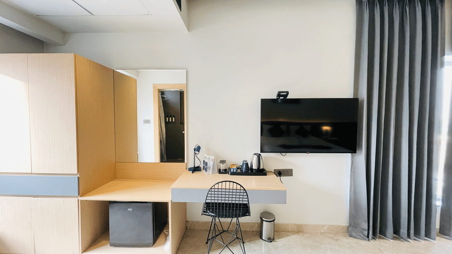 A hotel room at Hotel Kyzen HiTech City featuring a wooden work desk with chair wall-mounted TV grey curtains wooden cabinetry and a large mirror in a modern setting.