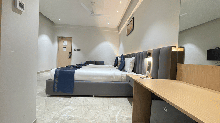 A twin bed room at Hotel Kyzen HiTech City featuring two beds with white bedding blue accent pillows grey headboards a wooden work desk and light tiled flooring in a modern layout.