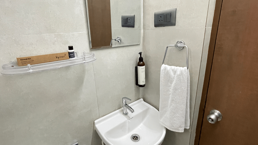 Bathroom at Hotel Kyzen Smart Stays Madhapur featuring a wall-mounted wash basin, mirror, toiletry bottles, towel holder, and grey tiled walls.