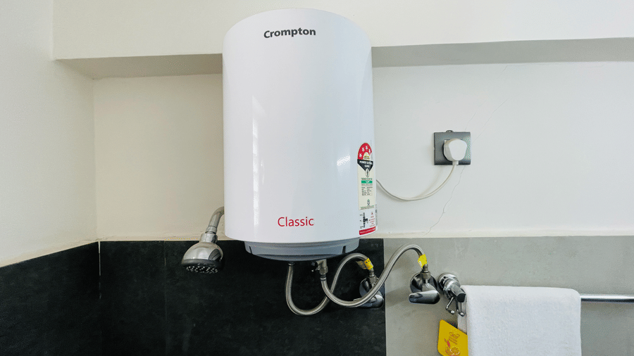 Bathroom corner at Hotel Kyzen Smart Stays Madhapur featuring a Crompton electric geyser, towel rail with folded towels, and dark tiled walls.