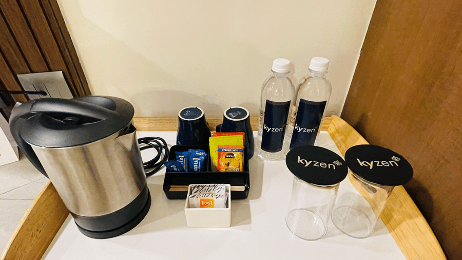 In-room amenity counter at Hotel Kyzen Smart Stays Madhapur featuring an electric kettle, water bottles, tea bags, and branded Kyzen toiletry pouches on a wooden counter.