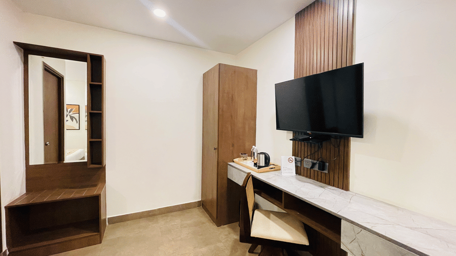 Corner view of a hotel room at Hotel Kyzen Smart Stays Madhapur featuring a wall-mounted TV, wooden wardrobe, dressing mirror, work desk, and warm ceiling lighting.