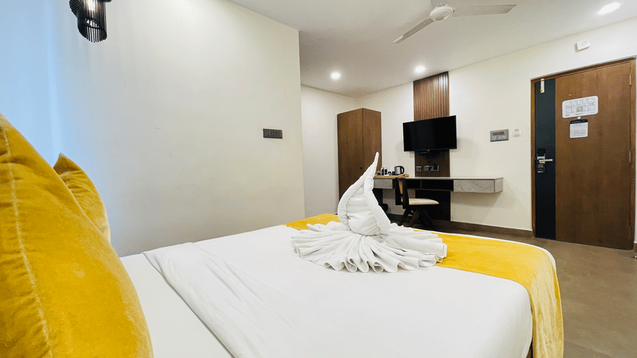 Close-up view of a hotel bed at Hotel Kyzen Smart Stays Madhapur with bright yellow pillows, white bedding, swan towel folding, and wooden furniture alongside.