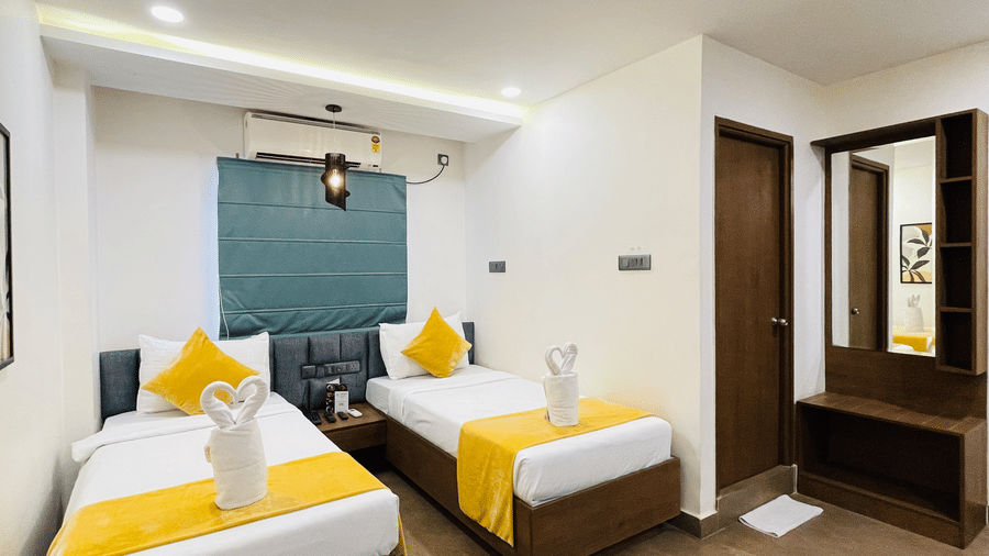 Full view of twin bed room at Hotel Kyzen Smart Stays Madhapur with yellow accent bedding, teal headboard panel, wooden furniture, and warm recessed ceiling lighting.