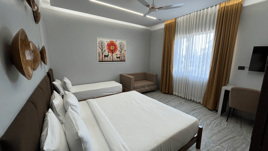 A wide-angle shot of the Suite featuring a double bed and a single bed facing towards a window covered with translucent white curtains at Hotel Mumbai House, Pithampur.