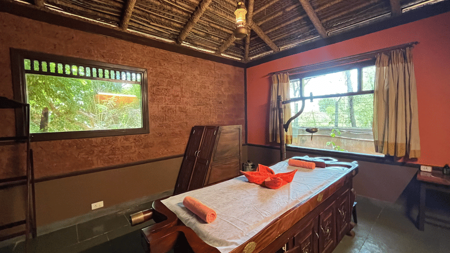 Well-lit Ayurveda spa room at Hoysala Village Resort featuring a spa bed with towels arranged on it under a thatched ceiling.