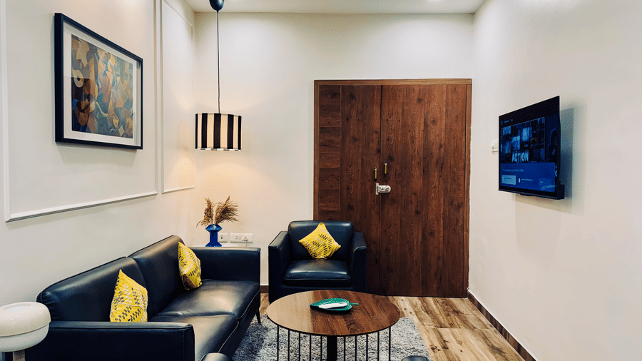 Side view of the living room of Coral 4BHK at Kove Smart Stays featuring upholstered seating areas facing a wall-mounted TV in a room with wooden flooring and ample lighting.