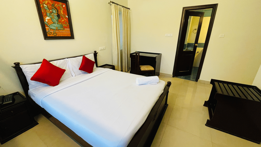 Hotel room with a double bed, red accent cushions, an air conditioner, and traditional wall art at Sanctum Suites Indiranagar, Bangalore.