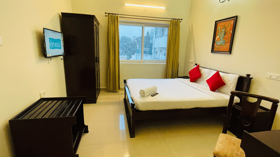 Hotel room with a double bed, red accent cushions, brown curtains, and a television at Sanctum Suites Indiranagar, Bangalore.