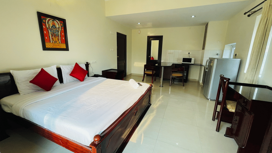 Spacious hotel room with a double bed, red accent cushions, traditional wall art, and a refrigerator at Sanctum Suites Indiranagar, Bangalore.