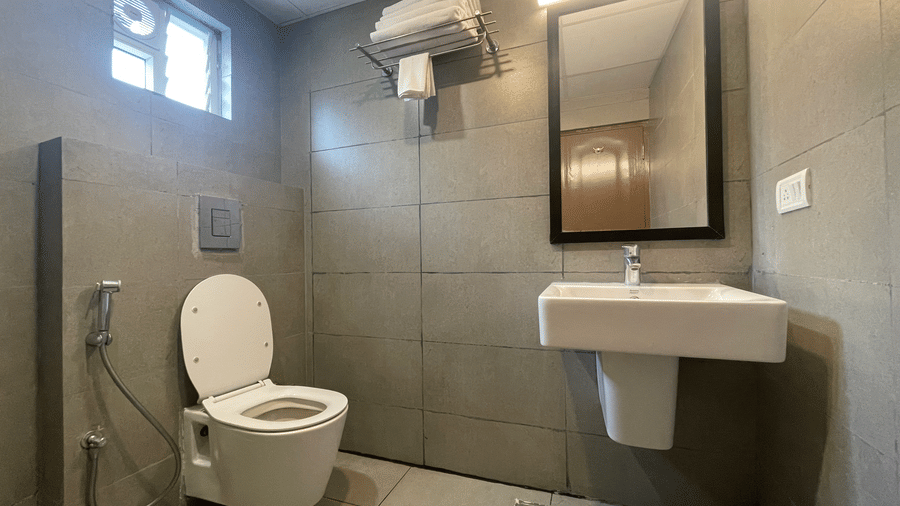 Bathroom at Sanctum Suites, Richmond Road, Bangalore has a toilet, a white sink, a mirror, and grey tiled wall with metal fixtures.