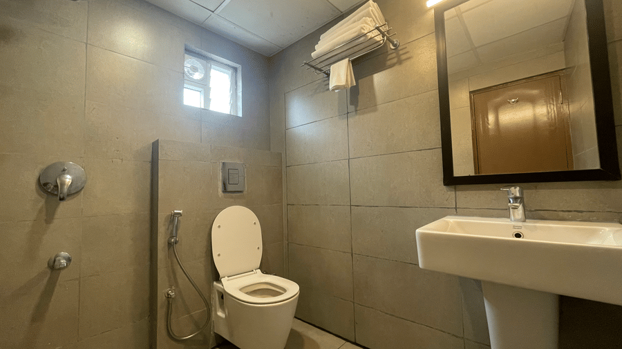 Bathroom at Sanctum Suites, Richmond Road, Bangalore features a toilet, a white washbasin, a mirror, and grey tiled walls with chrome fixtures.